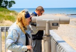 2 people standing on a boardwalk overlooking a beach. One person is peering through a telescope.